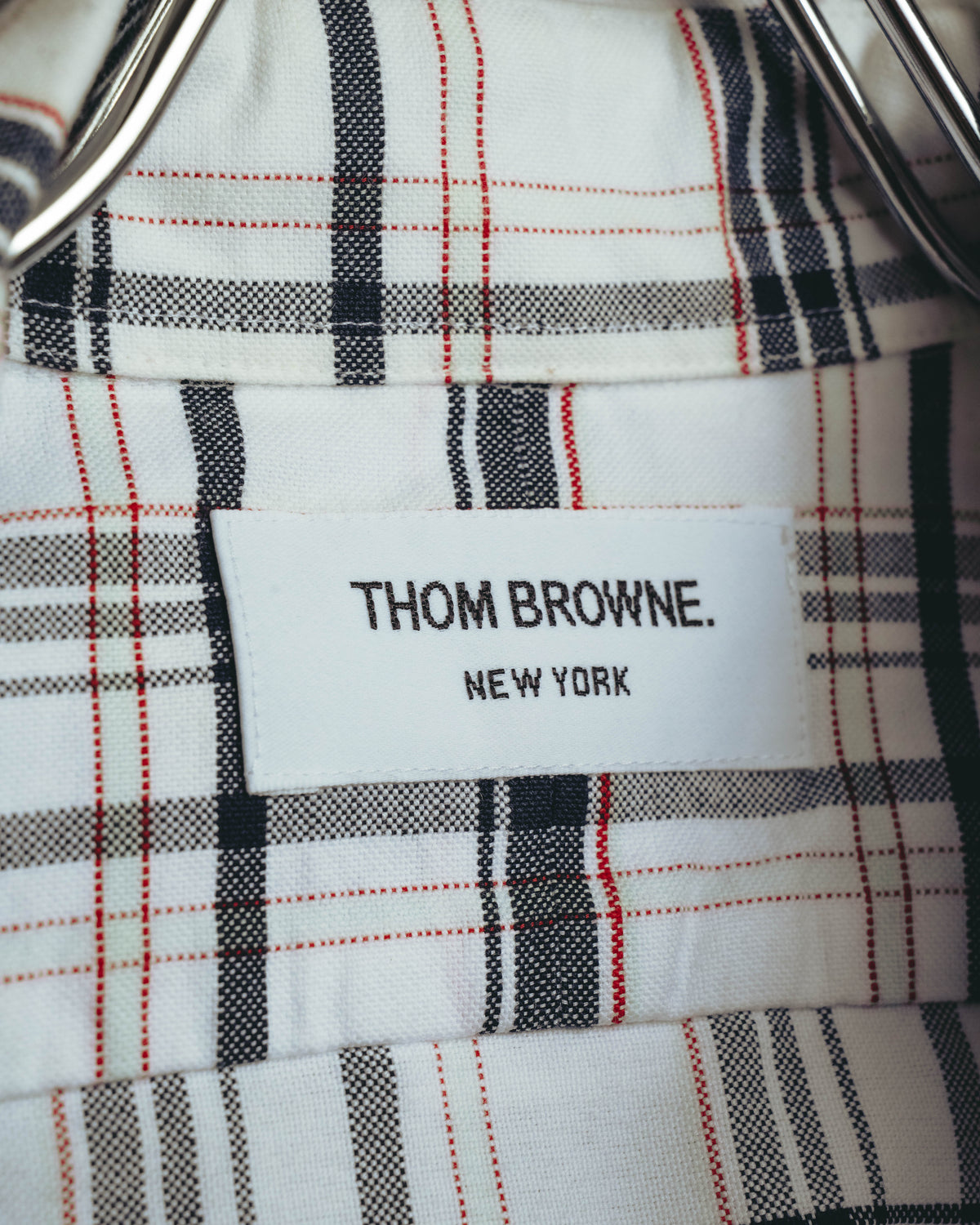 ホワイト、ブラック、グレーに細いレッドラインが交差するタータンチェック地に配された、「THOM BROWNE. NEW YORK」表記のネームタグ。