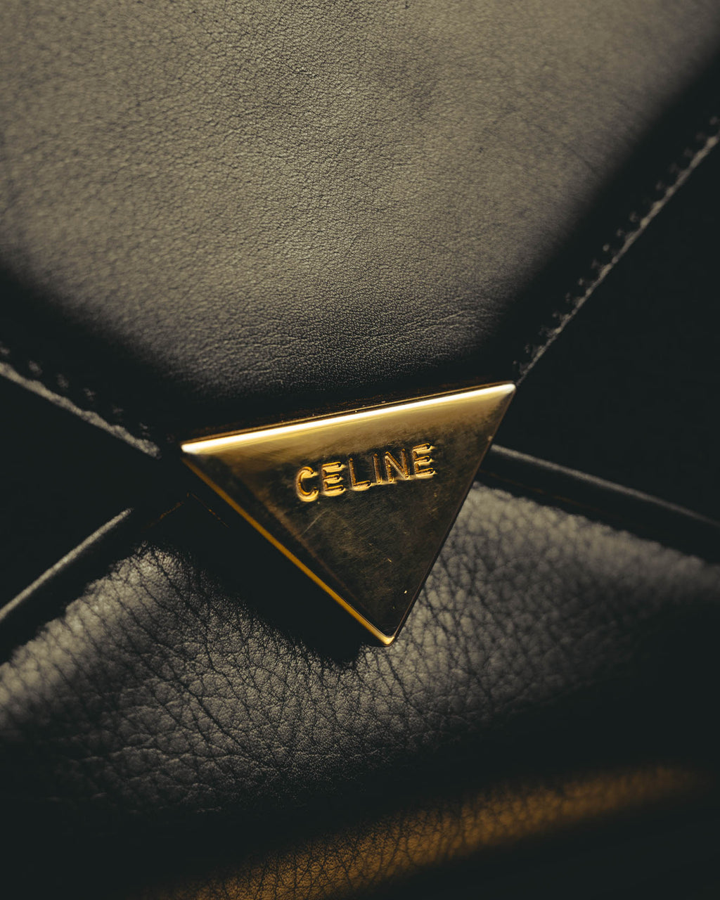 シャープな封筒型フラップに配された、CELINEの大文字刻印入り三角金具の留め具（クラスプ）を捉えたディテールカット。ブラックの表面とゴールドカラーの金具、フラップ周囲のステッチが確認できます。