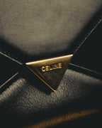 シャープな封筒型フラップに配された、CELINEの大文字刻印入り三角金具の留め具（クラスプ）を捉えたディテールカット。ブラックの表面とゴールドカラーの金具、フラップ周囲のステッチが確認できます。