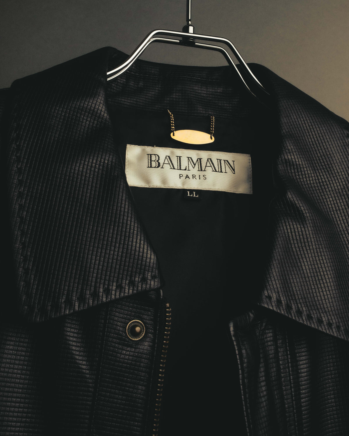 内側のBALMAIN PARISタグとLL表記、フロントのファスナーおよびスナップボタンを捉えた襟元のディテールカット。細かな格子状の型押しを施したブラックレザーの表情も確認できます。