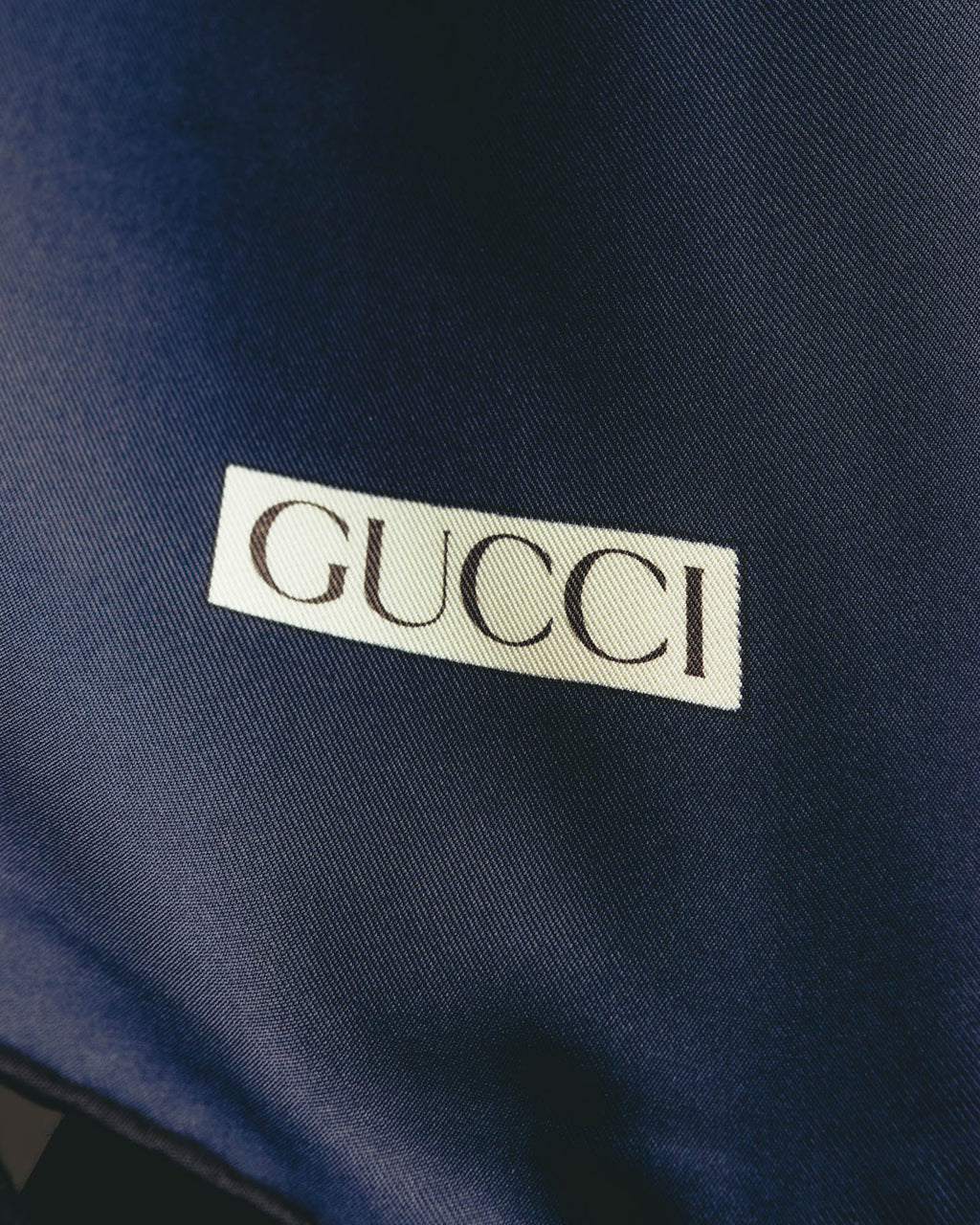 深いネイビーの生地に、クリーム色の枠とセリフ体のGUCCIロゴを配したディテールカット。細かな綾目を思わせるシルク生地の表情も確認できます。