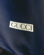 深いネイビーの生地に、クリーム色の枠とセリフ体のGUCCIロゴを配したディテールカット。細かな綾目を思わせるシルク生地の表情も確認できます。
