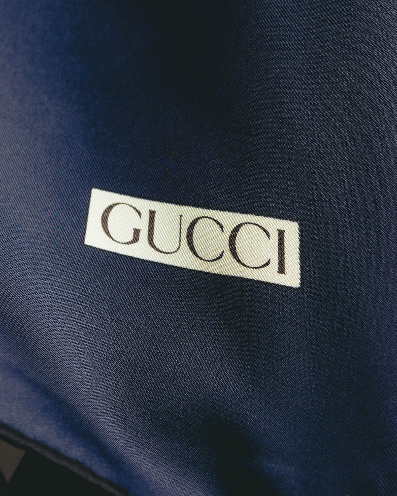 深いネイビーの生地に、クリーム色の枠とセリフ体のGUCCIロゴを配したディテールカット。細かな綾目を思わせるシルク生地の表情も確認できます。
