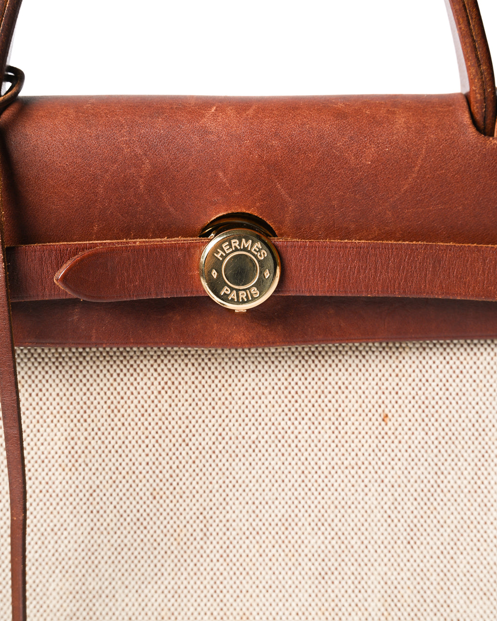 フロントのレザーベルトを通して留める、HERMÈS PARIS刻印入りゴールドカラーのセリエ金具を捉えたディテールカット。深いブラウンのレザートップパーツと、細かなブラウンの織りが見えるキャンバス調ボディの対比も確認できます。