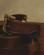 ゴールド調の透かし装飾バックルと、ダークブラウンの帯材の剣先を捉えたディテールカット。輪郭に沿うステッチとベルトループの仕立てが確認できます。