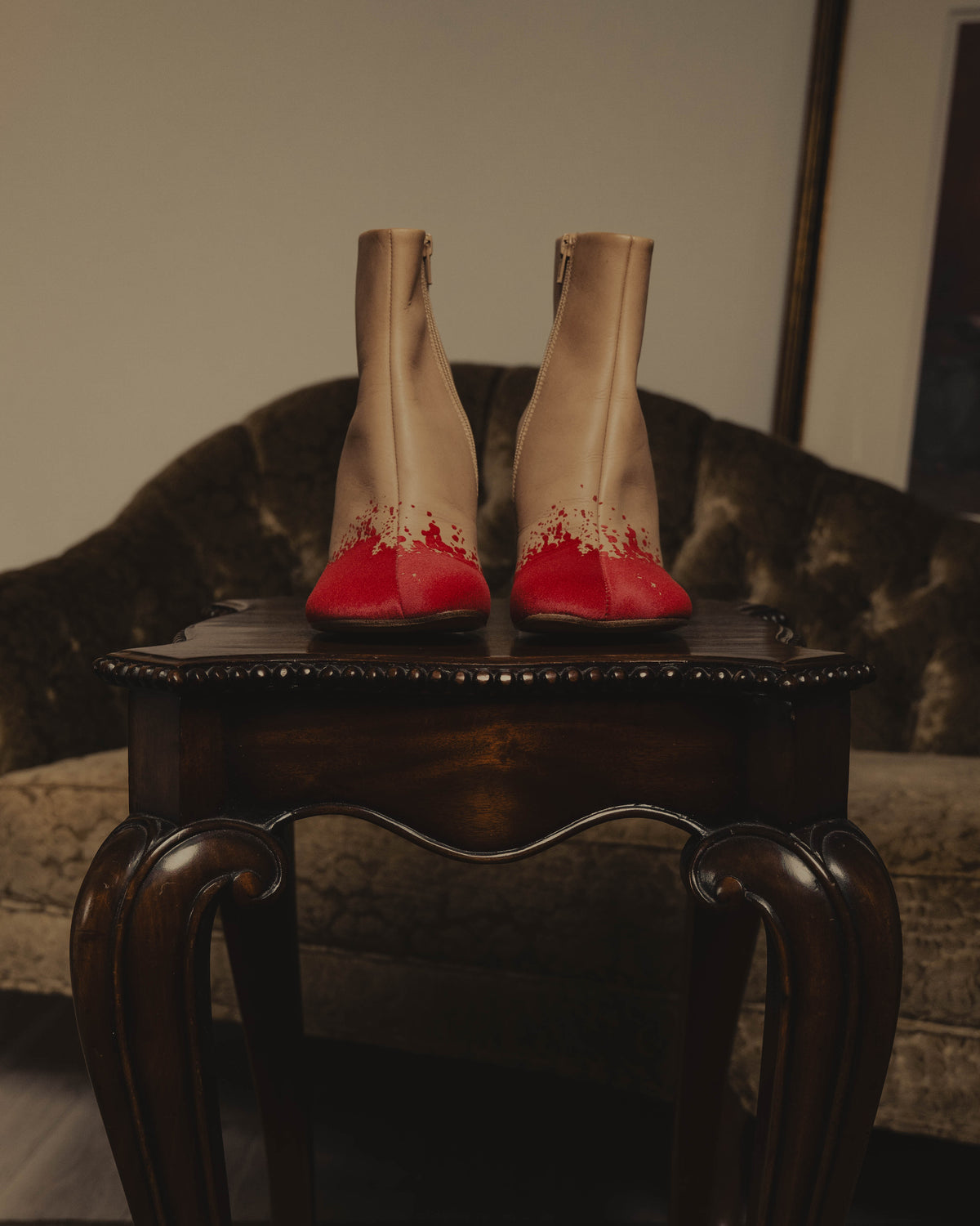 グレージュからベージュ系のレザーを基調に、つま先から甲へ鮮やかな赤の飛沫状ペイントが広がる正面全体像。丸みを帯びたアナトミックトウと、内側に配されたファスナーの輪郭が確認できます。