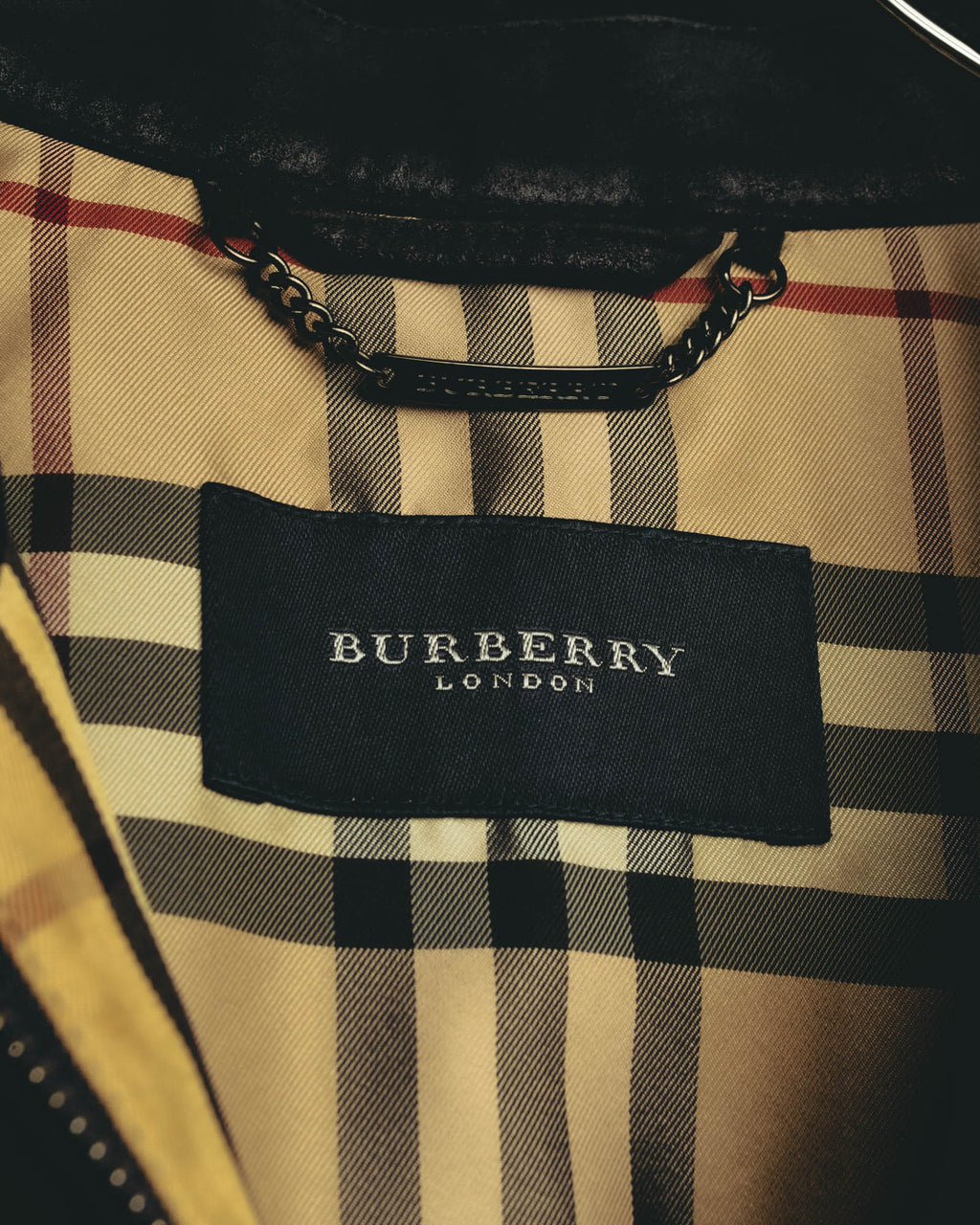 チェック柄の内側に配されたBURBERRY LONDONの織りタグと、襟裏のブランド刻印入り金属製吊りチェーンプレートを捉えたカット。ベージュを基調にブラック、ホワイト、レッド系のラインが走るチェック柄も確認できます。