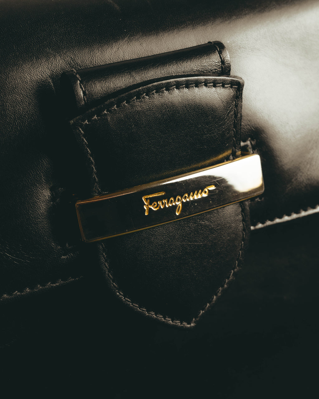 フロントに配された、Ferragamoのスクリプトロゴを刻んだゴールドカラーのプレートを捉えたディテールカット。ブラックレザーのフラップと周囲のステッチも確認できます。