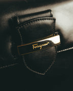 フロントに配された、Ferragamoのスクリプトロゴを刻んだゴールドカラーのプレートを捉えたディテールカット。ブラックレザーのフラップと周囲のステッチも確認できます。