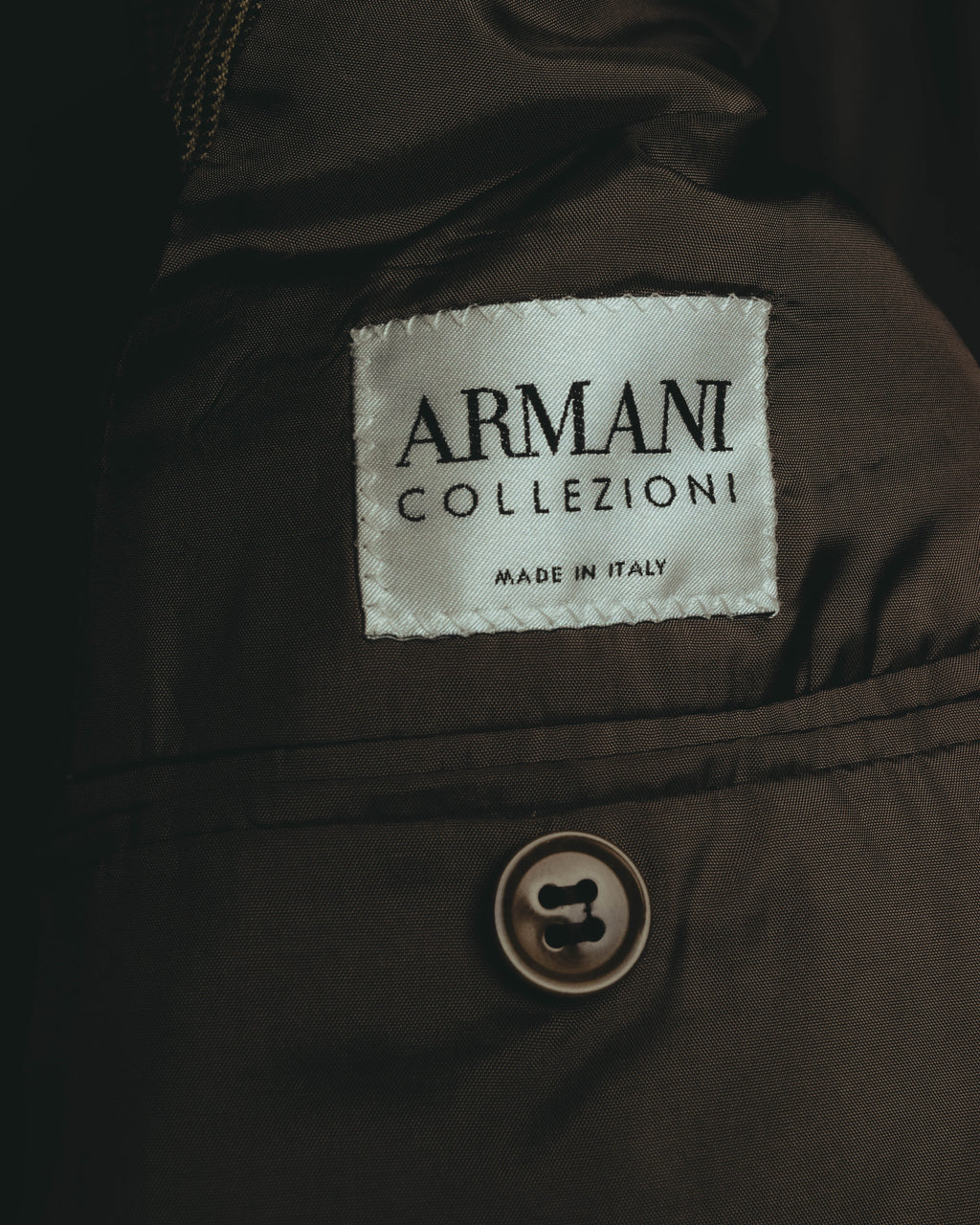 「ARMANI COLLEZIONI」とMADE IN ITALY表記が確認できる内タグのカット。落ち着いたブラウンの裏地と、全体の色彩に溶け込む同系色ボタンも写しています。