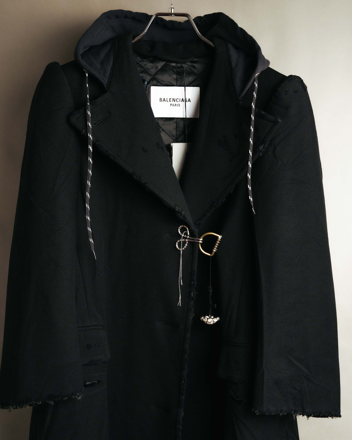 白地に黒文字で「BALENCIAGA PARIS」と記された襟元のタグを捉えたカット。一体化したチャコールブラック調のフード、キルティング仕立ての裏地、異なる金属パーツを組み合わせたフロントの留め具も確認できます。