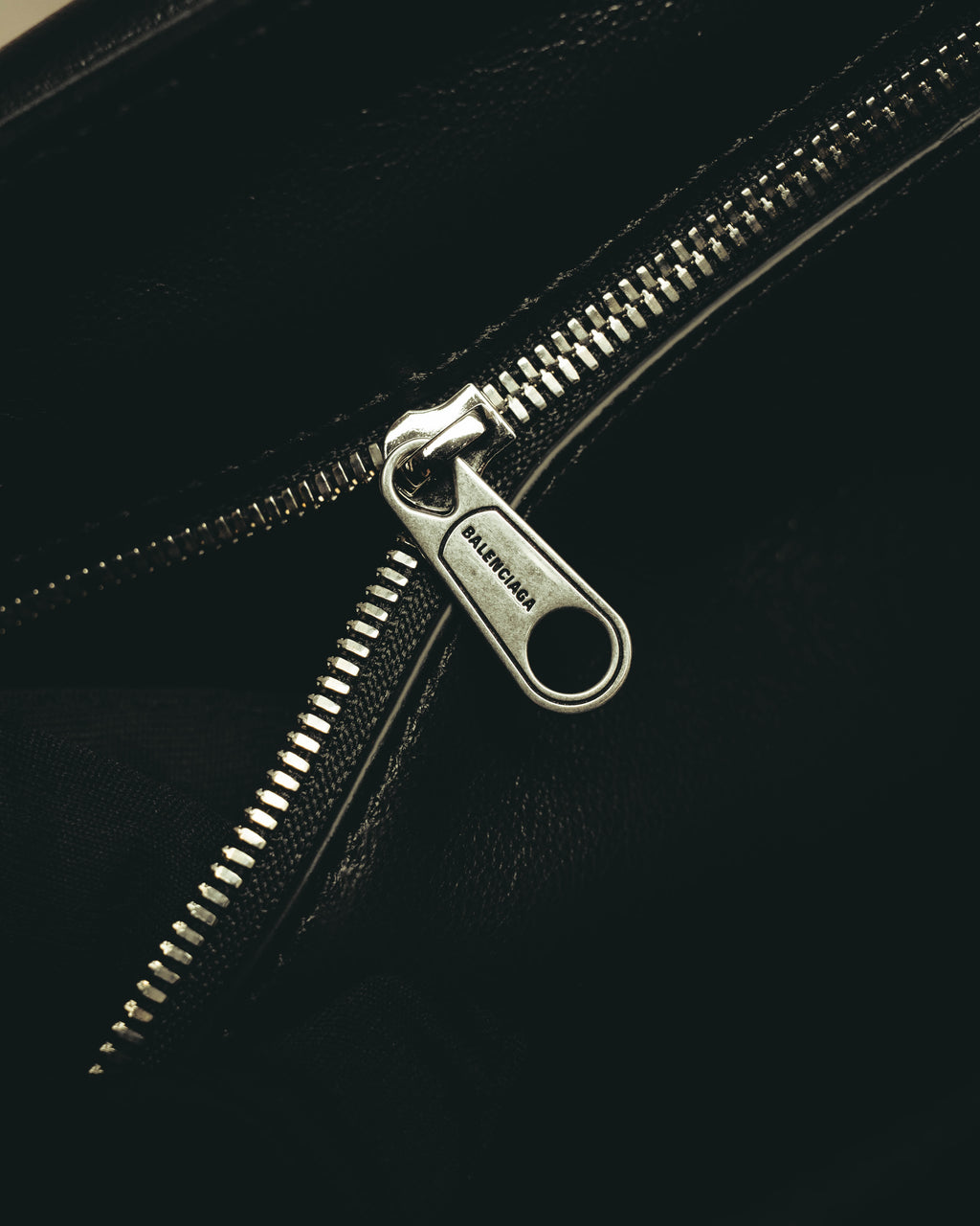 内装のファスナーポケットに配された、BALENCIAGA刻印入りシルバーカラーのジッパー引手を捉えたディテールカット。シボ感のあるブラック素材とファスナーの仕様が確認できます。