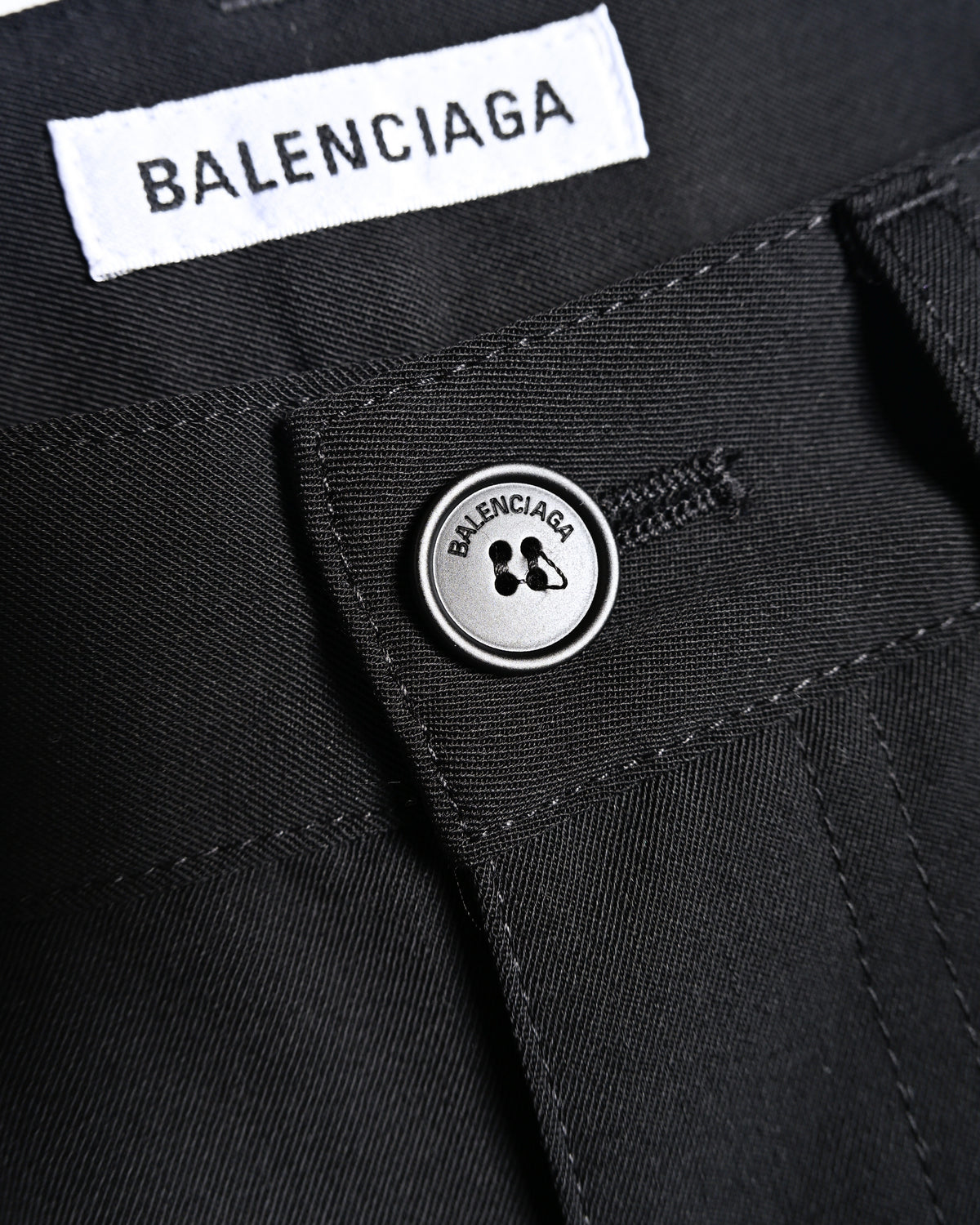 フロントのブランド名刻印入りオリジナルトップボタンと、ウエスト内側に配した「BALENCIAGA」ホワイトタグを捉えたディテールカット。ブラックのウールギャバジン調生地とウエスト周りの縫製も確認できます。