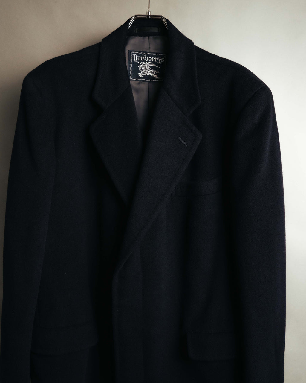 襟元に配された騎馬騎士図案入りの旧Burberrysネームと、端正なノッチドラペルを捉えたディテールカット。ダークネイビー系の表地とキュプラ・ポリエステル裏地も確認できます。