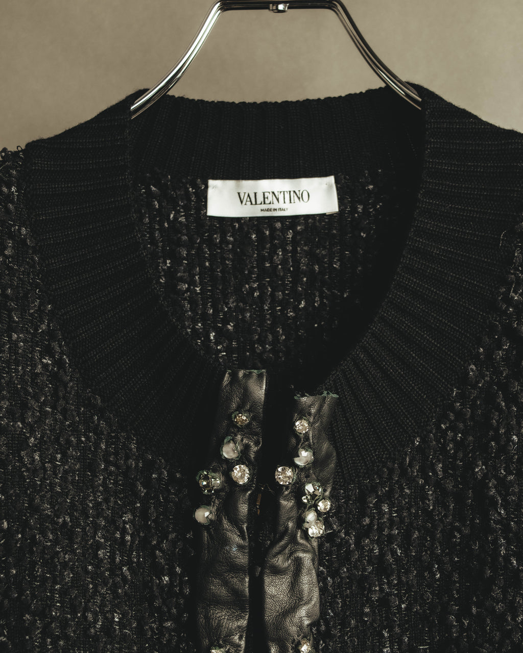 襟元に付く「VALENTINO MADE IN ITALY」のネームタグと、クリアストーンを爪留め状に配したブラックレザーの前立て。ループ状の凹凸を持つブラックの編地も確認できるカット。