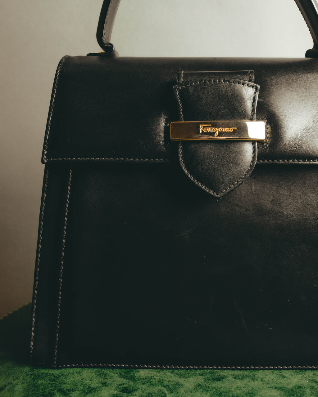 フロントフラップに配された、Ferragamoのスクリプトロゴ入りゴールドカラープレートのディテール。ブラックのスムースレザーと留め具周りのステッチを確認できるカットです。