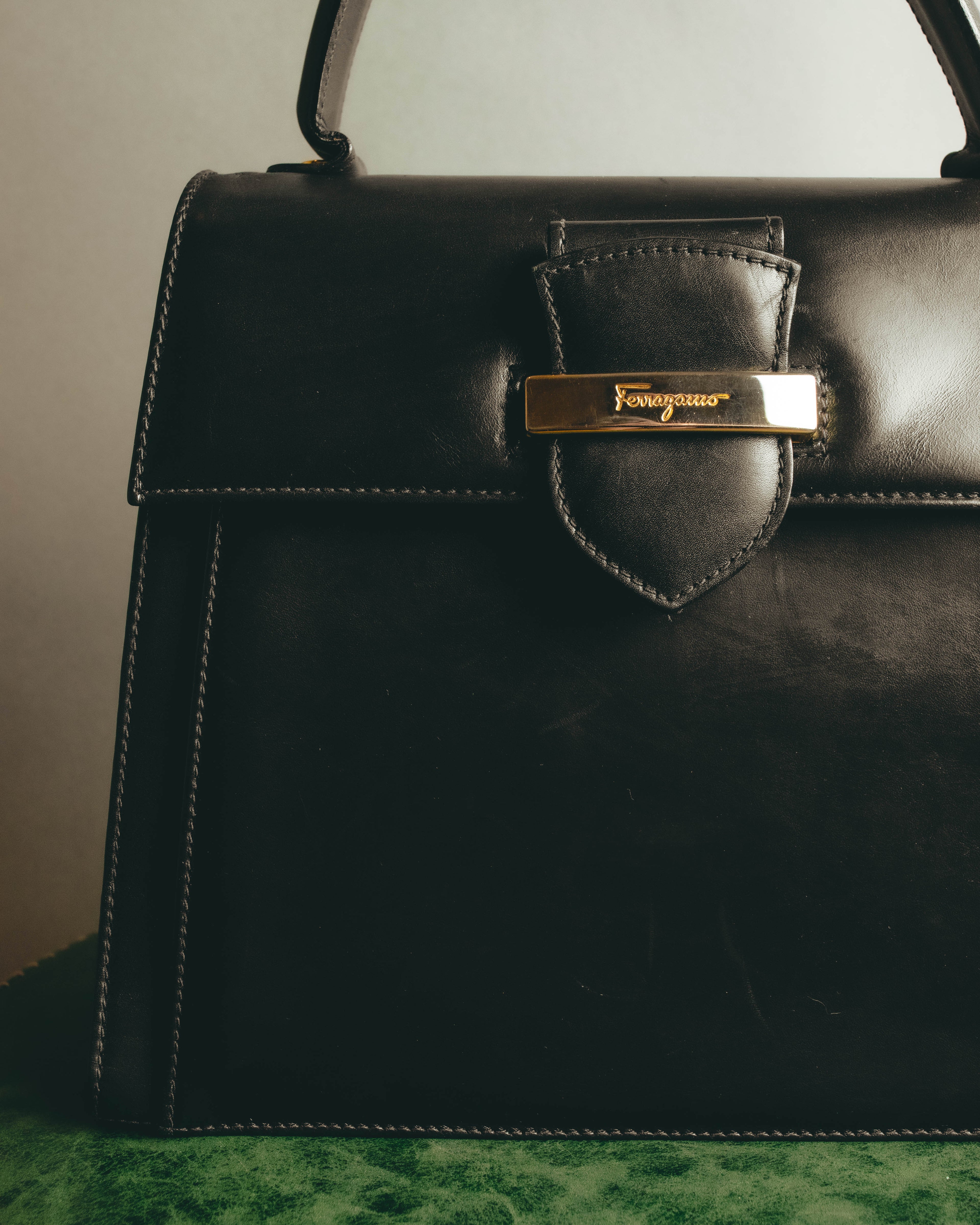 フロントフラップに配された、Ferragamoのスクリプトロゴ入りゴールドカラープレートのディテール。ブラックのスムースレザーと留め具周りのステッチを確認できるカットです。