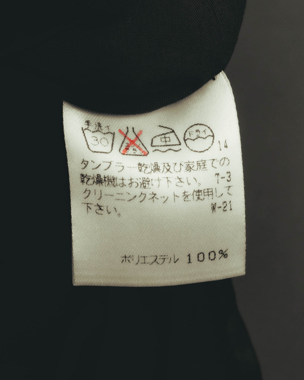 旧JIS方式の洗濯表示と、ポリエステル100%の素材表記を確認できる品質表示タグ。タンブラー乾燥禁止およびクリーニングネット使用のケア表記が記されています。