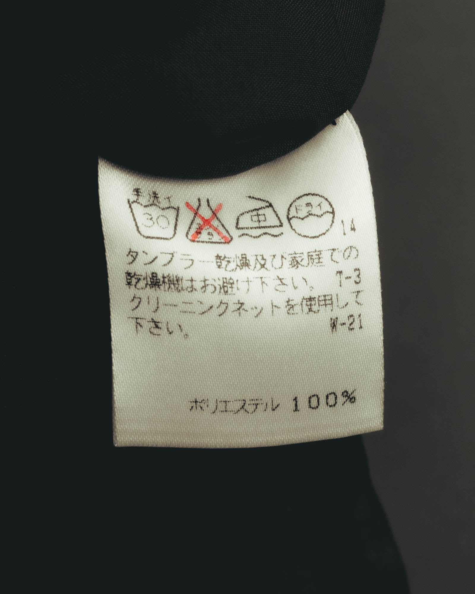 旧JIS方式の洗濯表示と、ポリエステル100%の素材表記を確認できる品質表示タグ。タンブラー乾燥禁止およびクリーニングネット使用のケア表記が記されています。
