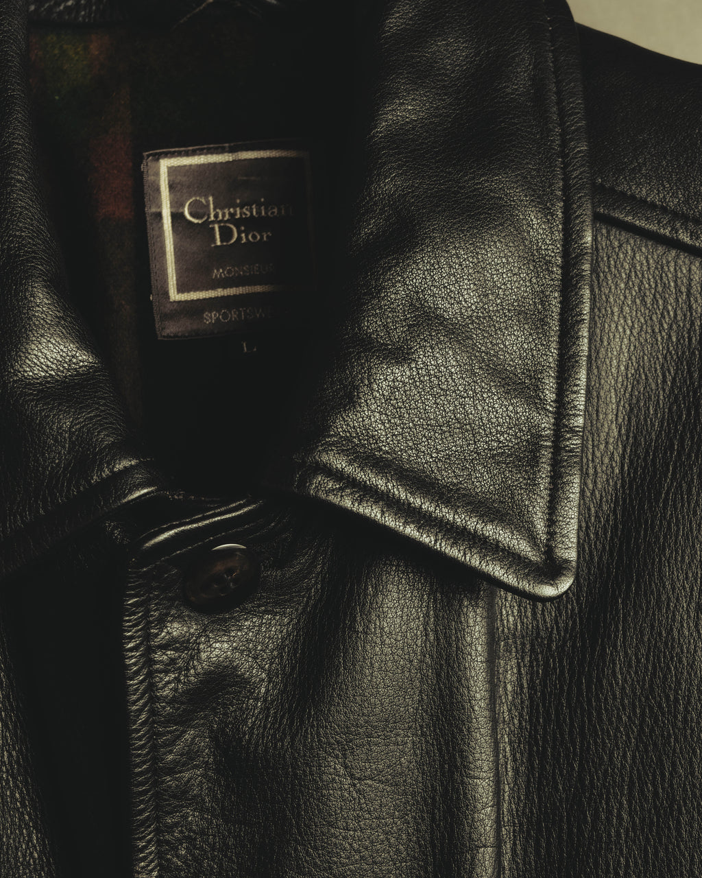 襟元に配された Christian Dior Monsieur Sportswear の織りネームとLサイズ表記を捉えたディテールカット。粒状のシボを持つブラックレザー、大ぶりな襟、フロントの4穴ボタンも確認できます。