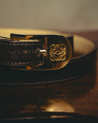 片側を緩やかに丸めた縦長フォルムのバックルに、四つのLを組み合わせたアナグラムが立体的に刻まれたディテールカット。鏡面に近い滑らかな金属面と、ブラックレザーの細やかなステッチを確認できます。