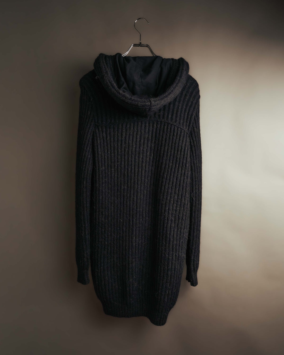 大型のフードからロング丈の身頃へと続く、コート背面の全体像。厚みのある畦編みと、縦方向に走る立体的な編み地が確認できるカット。