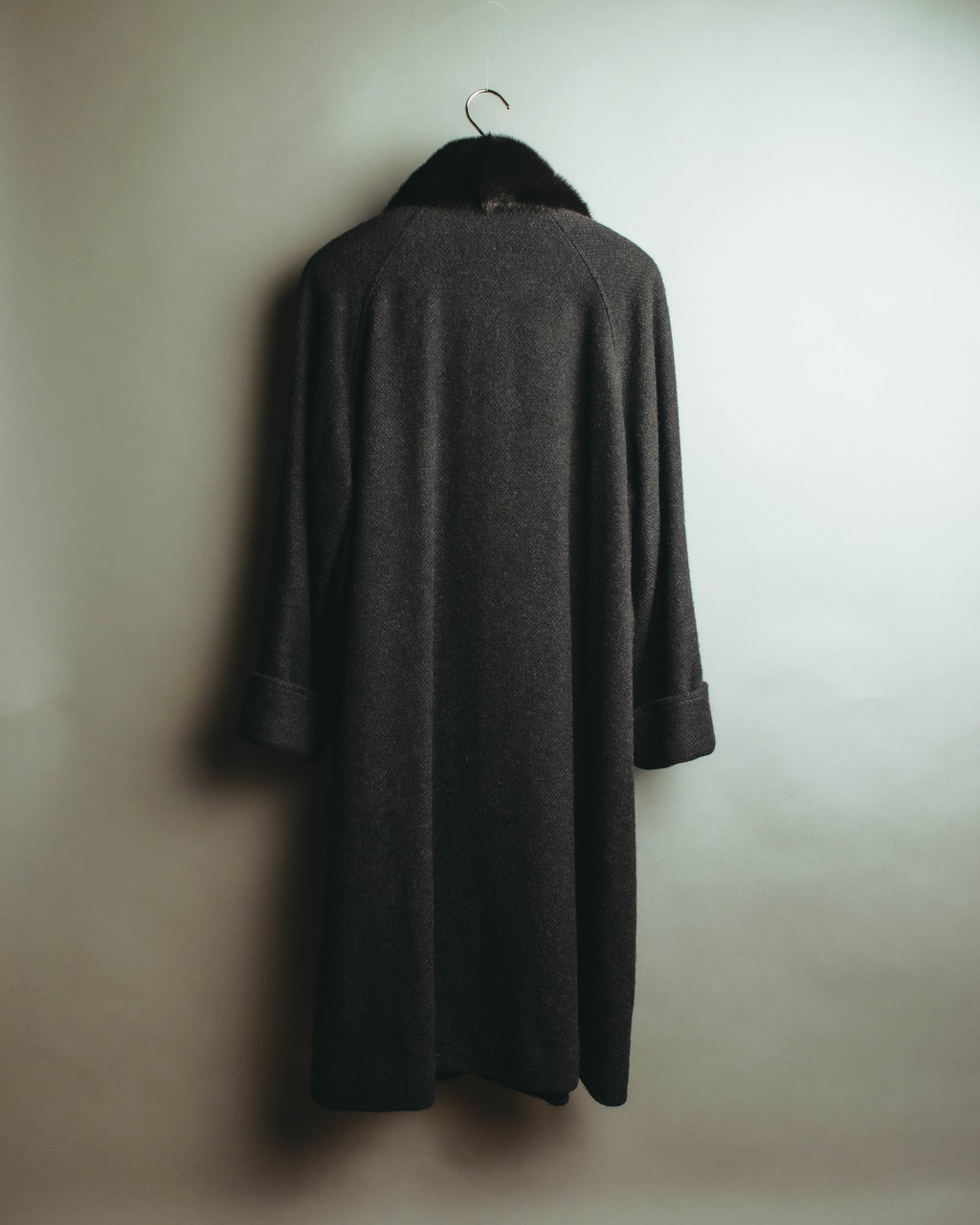 黒に近い深いチャコールグレーのロングコートを背面から捉えた全体像。なだらかな肩周りから裾へほぼ真っすぐに落ちる輪郭と、首元の毛足豊かなカラー、幅広く折り返された袖口が確認できます。