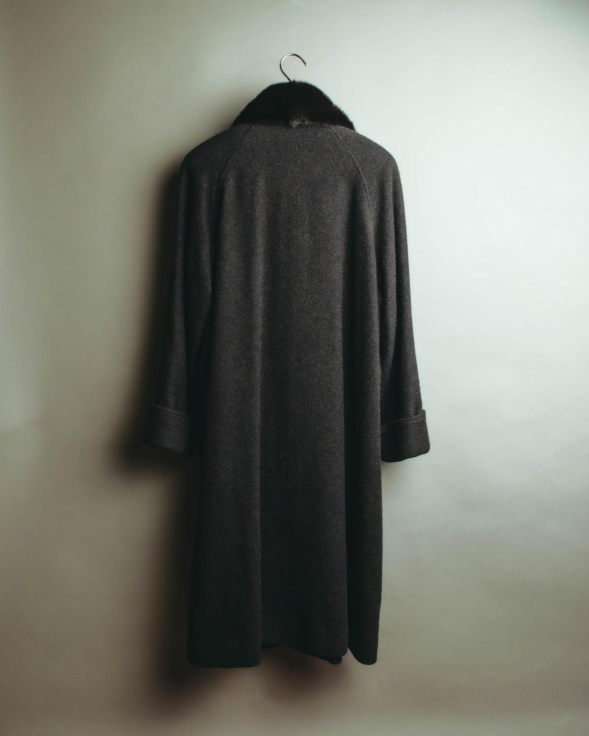 黒に近い深いチャコールグレーのロングコートを背面から捉えた全体像。なだらかな肩周りから裾へほぼ真っすぐに落ちる輪郭と、首元の毛足豊かなカラー、幅広く折り返された袖口が確認できます。