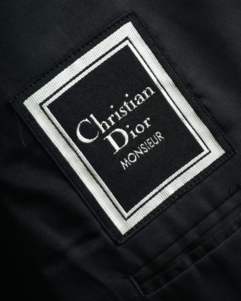 黒地に白い織り文字と二重枠を配した、Christian Dior Monsieurのブランドタグ。滑らかな裏地に縫い付けられた内側のタグを確認できるカット。