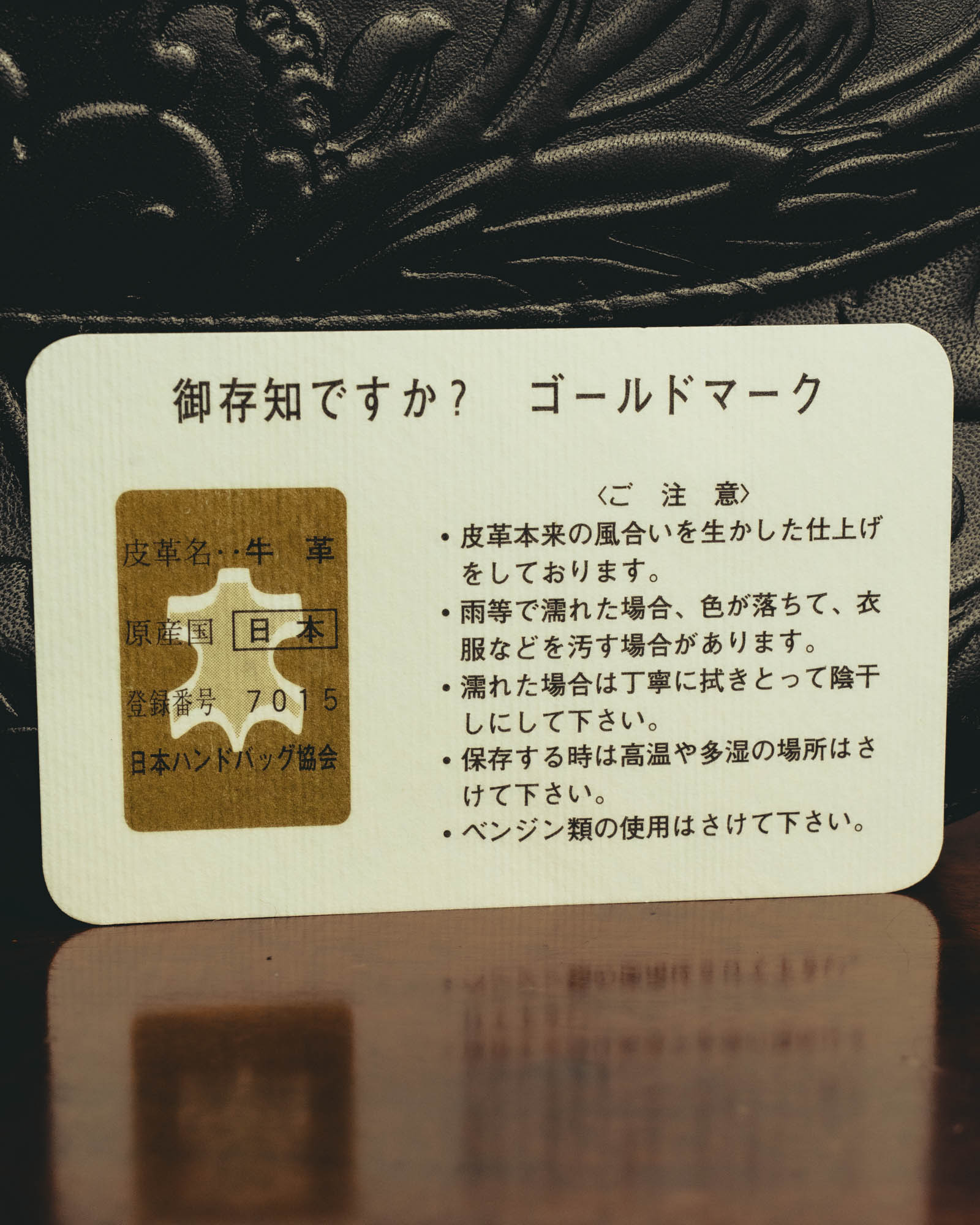 牛革、原産国日本、登録番号7015、日本ハンドバッグ協会の記載が確認できる付属カード。背景には立体的な型押しを施したブラックレザーが写ります。