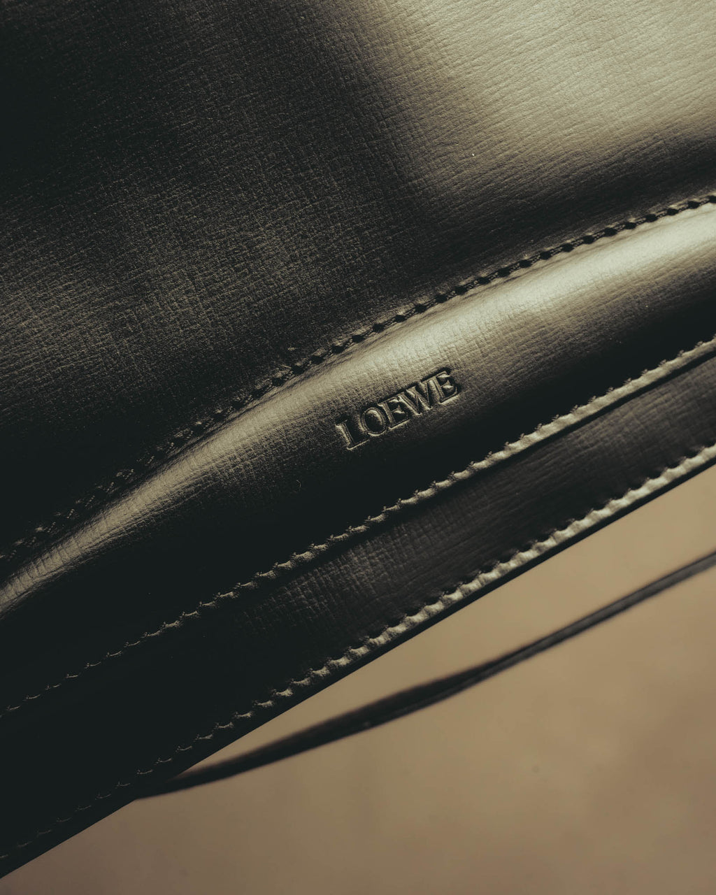 フラップ下に配された旧セリフ体のLOEWEロゴ刻印と、輪郭に沿って施されたステッチを捉えたディテールカット。艶を帯びたブラックの滑らかな表面も確認できます。
