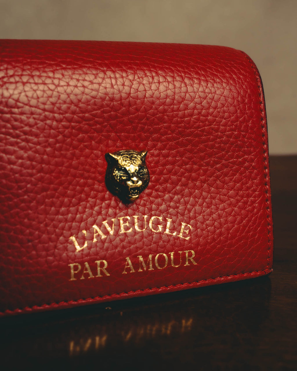 フロント中央に配された、ネコ科の動物を象ったアンティークゴールド調メタルパーツと「L'AVEUGLE PAR AMOUR」のゴールドレタリングのアップ。深みのあるレッドのグレインレザーと同系色ステッチも確認できるディテールカットです。