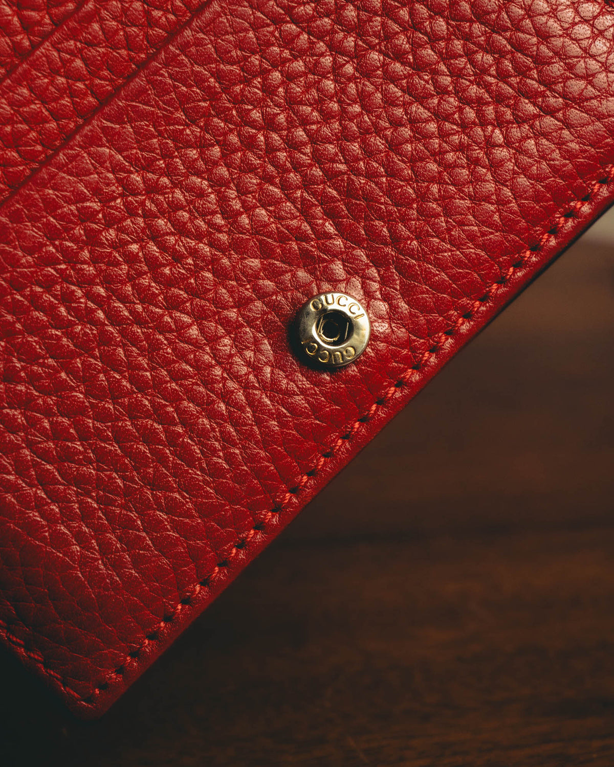 横長二つ折り構造の留め具となる、GUCCI刻印入りスナップボタンのディテールカット。細かなシボを持つレッドレザーと同系色のステッチ、ブラックのコバを確認できます。