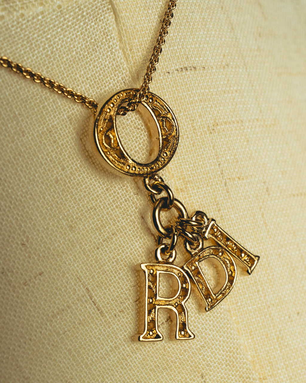 透かし模様と粒状のレリーフを施した円環状のOから、独立したD・I・Rの文字チャームが揺れるペンダントトップのディテール。ゴールドカラーのツイストチェーンを合わせた構成が確認できます。