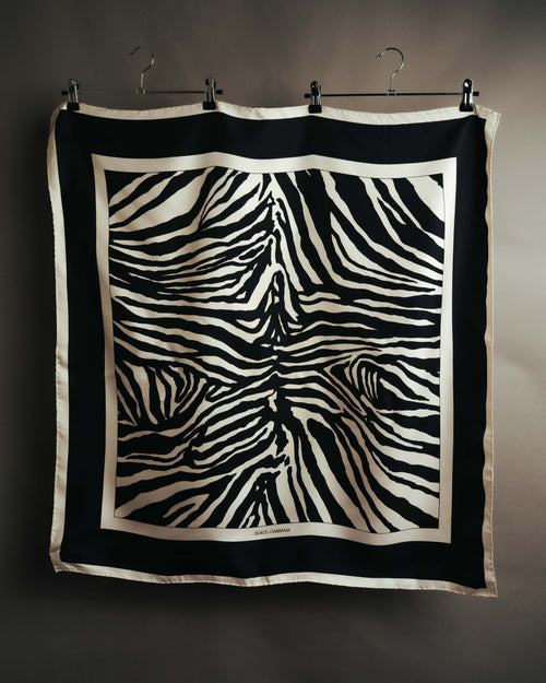 中央へ向かって広がる不規則なゼブラ柄と、外周の太いブラックボーダーを捉えた全体カット。四辺を細幅で処理したスクエア型の構造と、下部ボーダーに配されたロゴが確認できます。