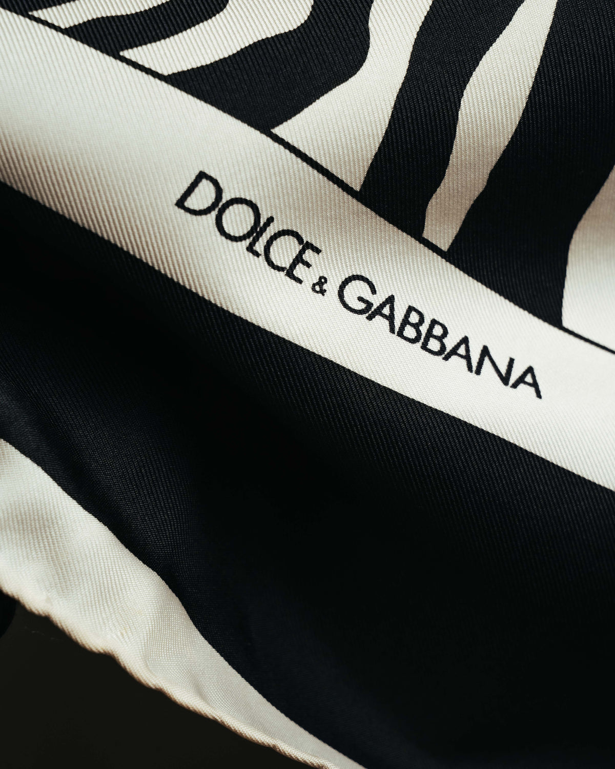 アイボリー系の額縁状ボーダーに配された DOLCE & GABBANA ロゴのディテール。ゼブラ柄と細かな斜文組織のツイル調生地も確認できるカットです。