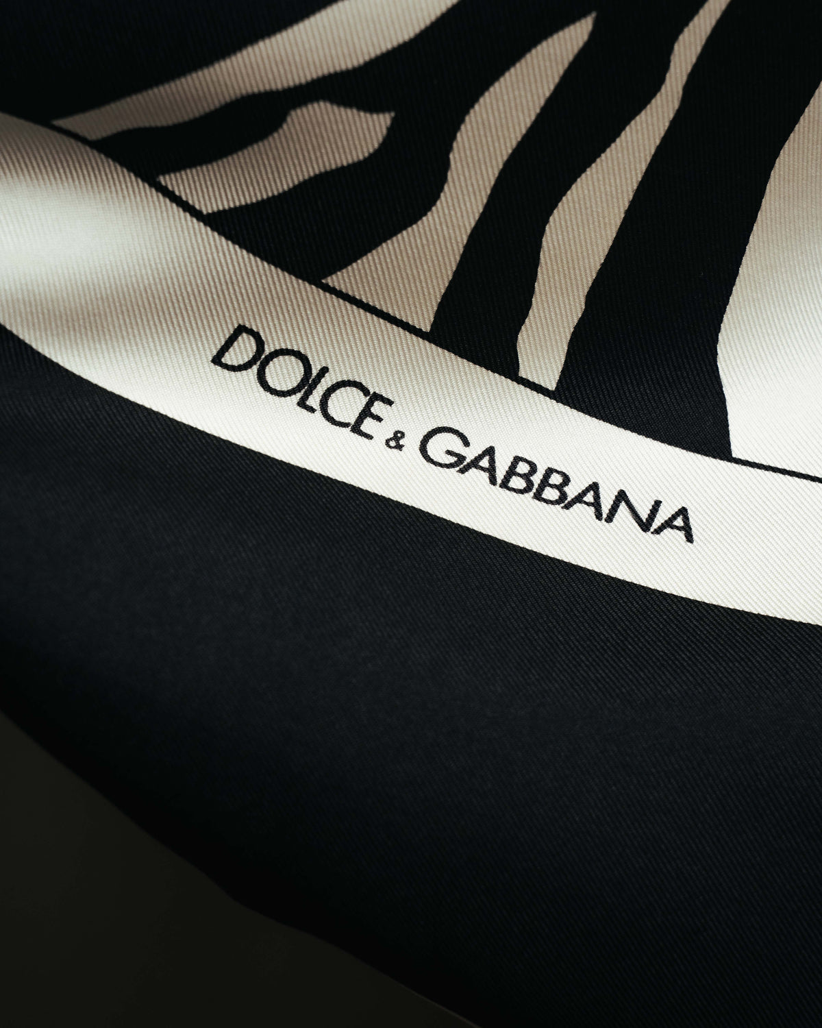 ゼブラ柄の外周を囲むアイボリー系ボーダーに、DOLCE & GABBANAのロゴが組み込まれたディテールカット。細かな斜文組織によるツイル調の生地感も確認できます。