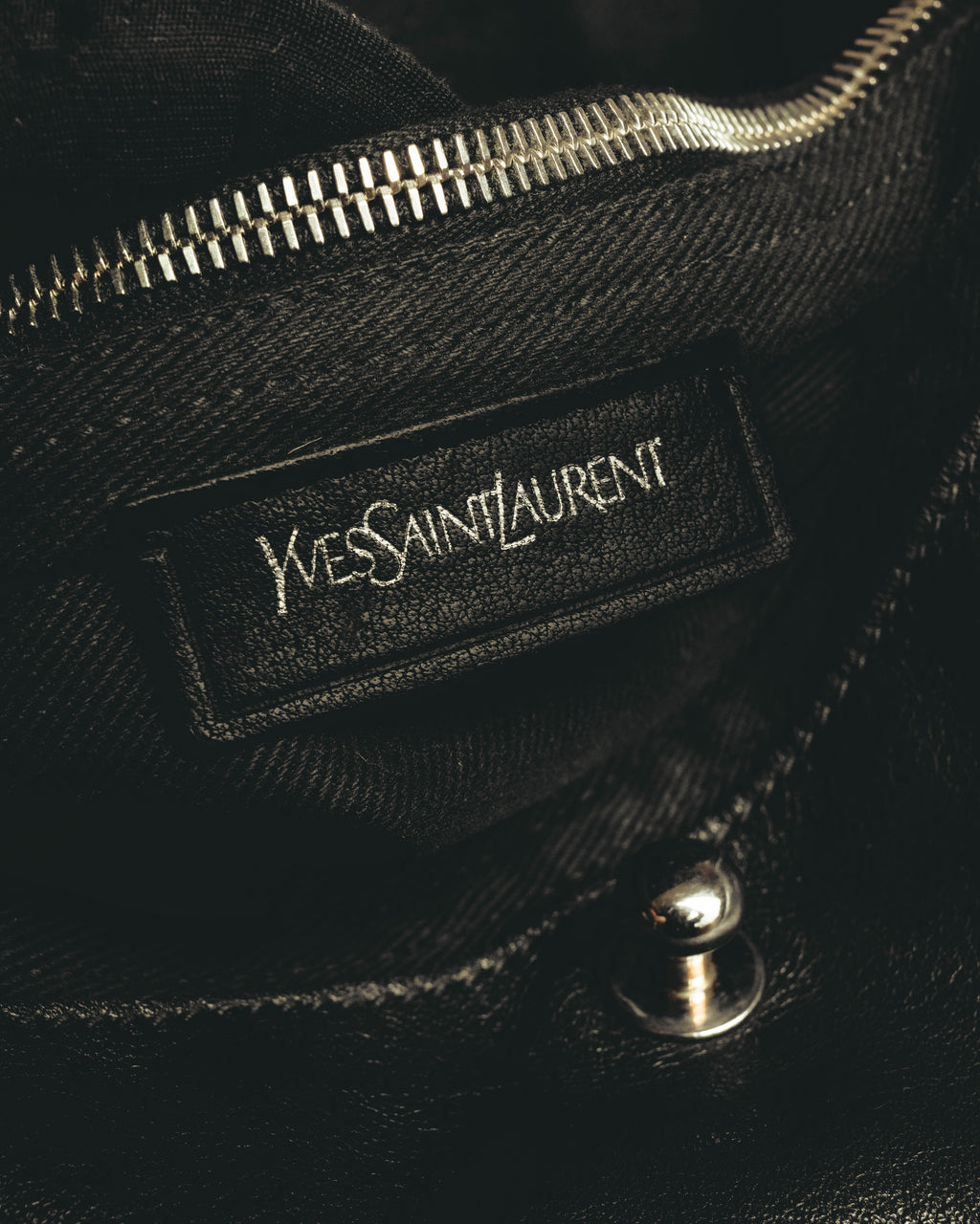 内側に配された旧 Yves Saint Laurent 表記のレザーパッチと、上部の内ポケットジッパーを捉えたカット。黒い内装とシルバーカラーのジッパーが確認できます。