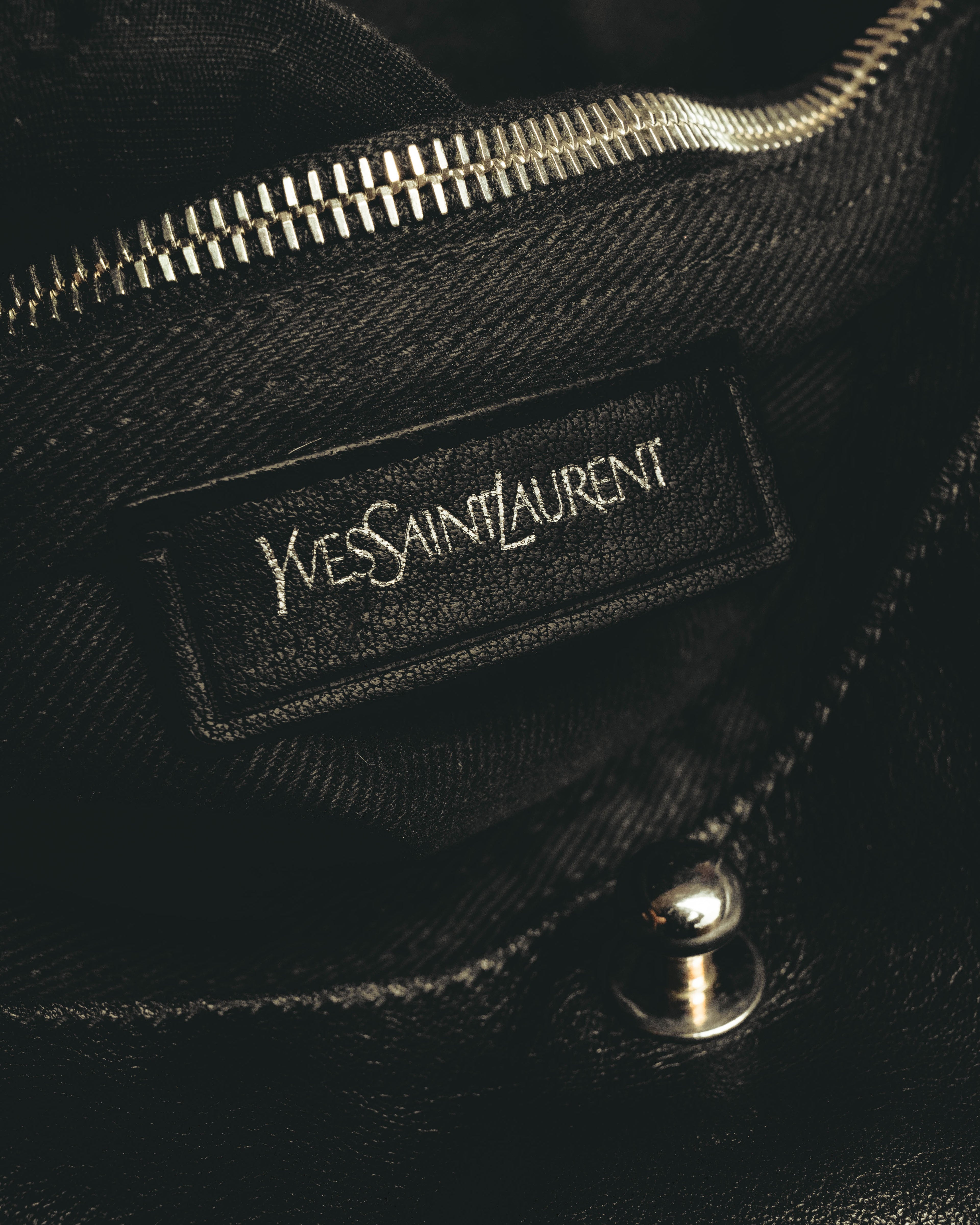 内側に配された旧 Yves Saint Laurent 表記のレザーパッチと、上部の内ポケットジッパーを捉えたカット。黒い内装とシルバーカラーのジッパーが確認できます。