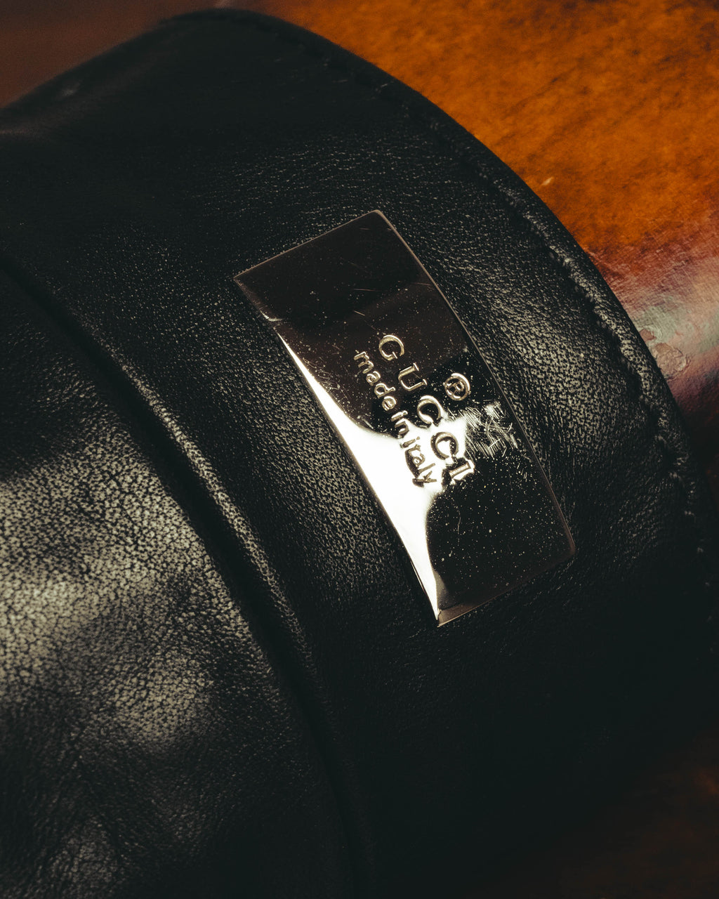 ブラックレザーの立体的なジップポケット中央に配した、GUCCI と made in Italy の刻印入り銀色メタルプレートのアップ。きめ細かなシボと落ち着いた艶を備えた本体とのコントラストが確認できます。