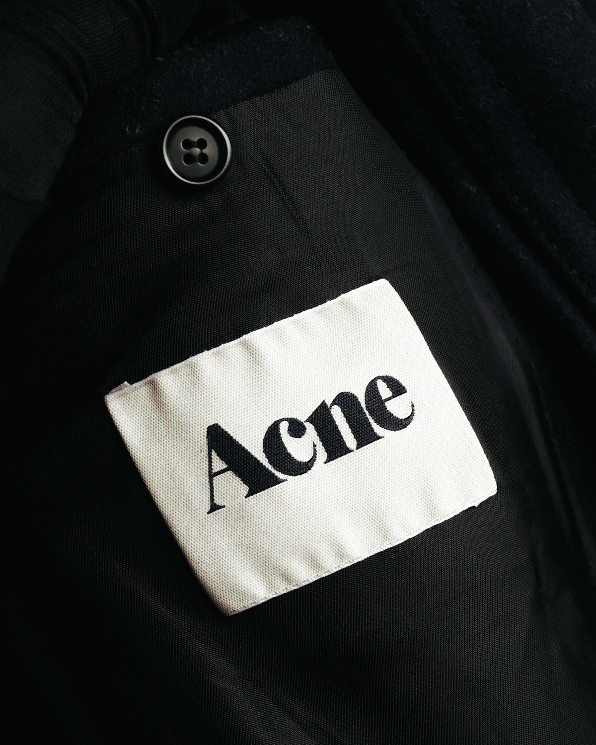 総裏仕立ての内側に備わる、生成り地に「Acne」表記を配したブランドタグのクローズアップ。旧ブランド表記を確認できる内タグと、隣接する裏地の四つ穴ボタンを捉えたカットです。