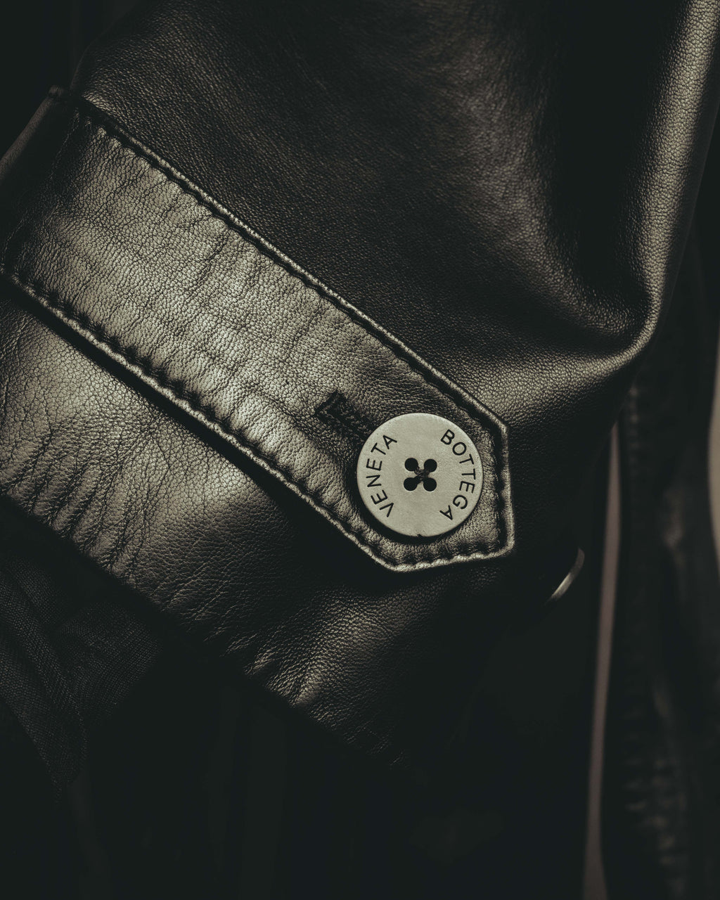 ブラックのリアルレザーを用いた袖口ストラップと、BOTTEGA VENETAの刻印が入る4穴ボタンを捉えたディテールカット。きめ細かなレザーの質感と縁のステッチも確認できます。