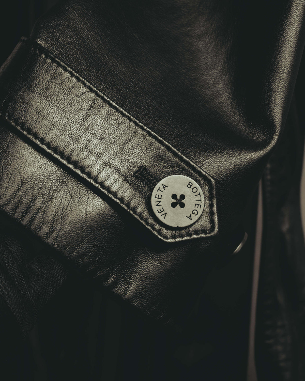 ブラックのリアルレザーを用いた袖口ストラップと、BOTTEGA VENETAの刻印が入る4穴ボタンを捉えたディテールカット。きめ細かなレザーの質感と縁のステッチも確認できます。