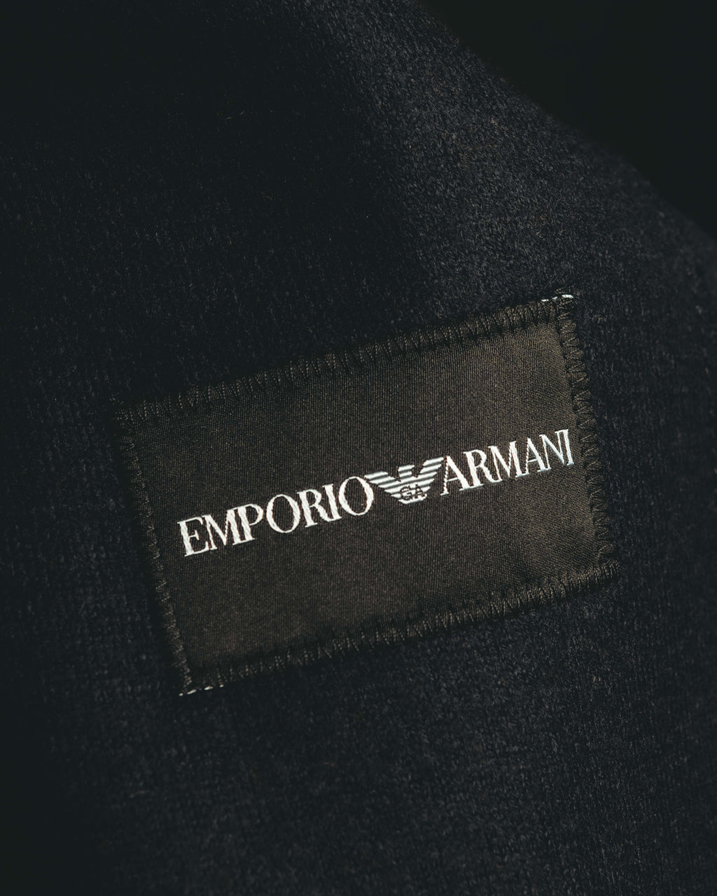 首元に配された、EMPORIO ARMANIの文字と白いイーグルロゴを記した黒地のブランドラベル。周囲には細やかな起毛感を備えた濃色の表地が写る。