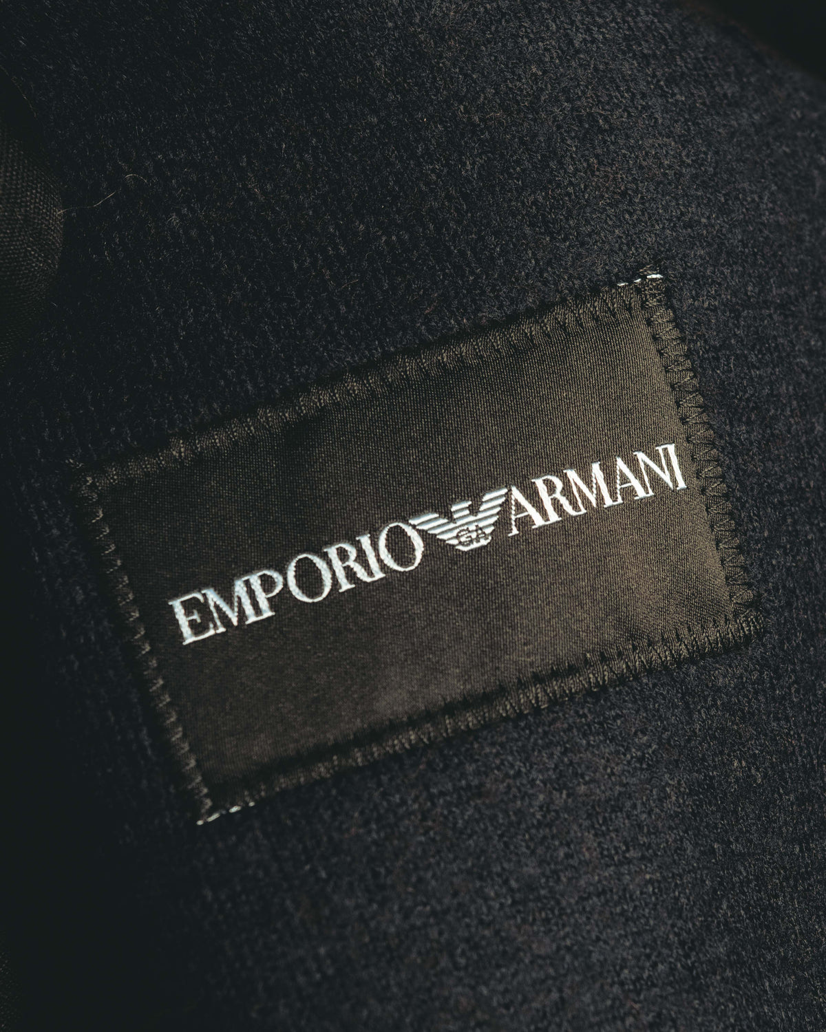 首元に配された、白いイーグルロゴとEMPORIO ARMANIの文字を記した黒地のブランドラベル。周囲には細やかな起毛感を備えた濃色の表地が写ります。