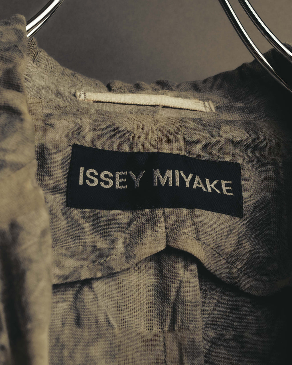 黒地に白いサンセリフ体で「ISSEY MIYAKE」を織り込んだネックタグのクローズアップ。周囲には顔料染めによる不均一な濃淡と皺の表情が見られます。