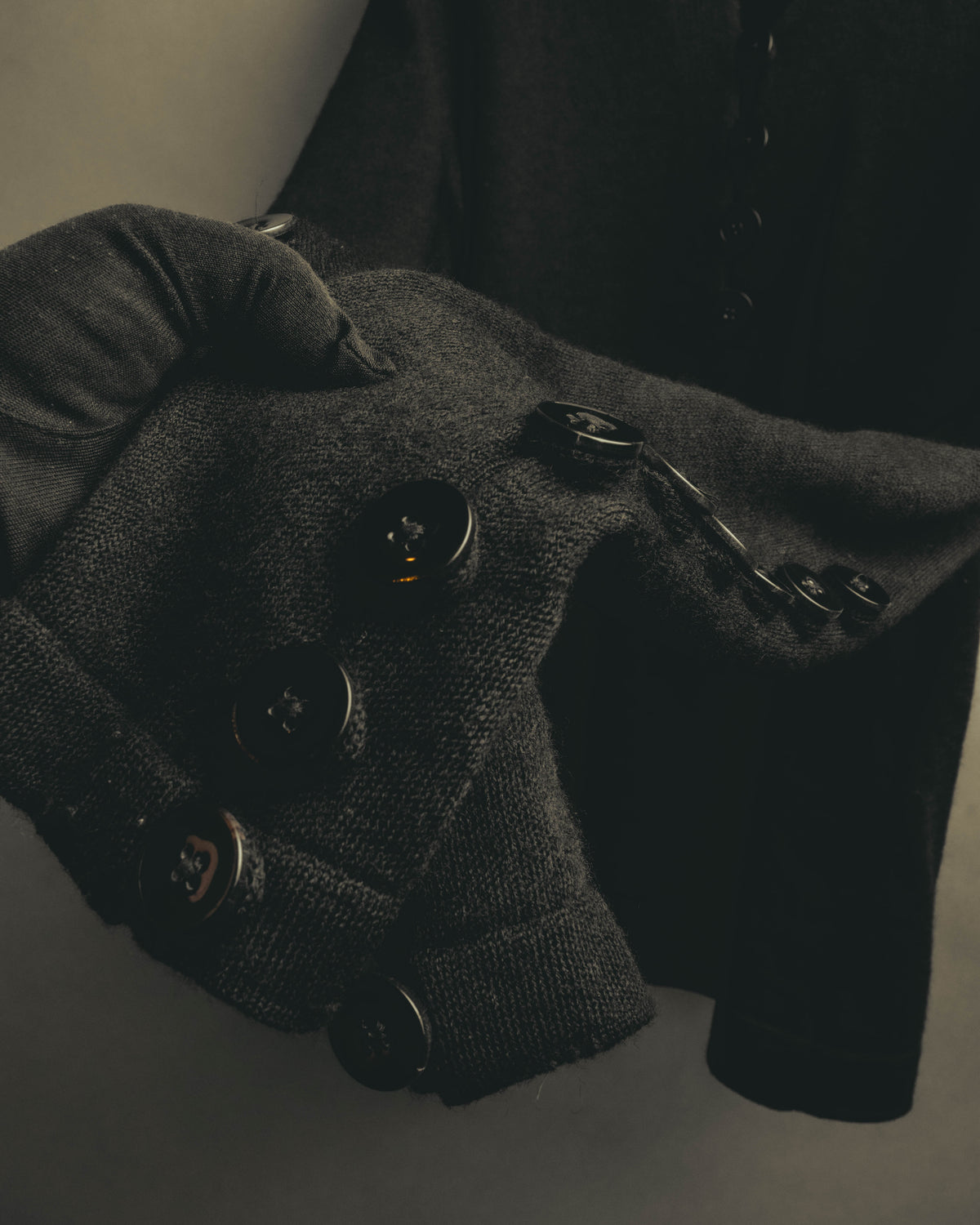 ブラックのニット地に、袖口から袖の内側へ複数のブラックボタンを連続して配したディテールカット。ボタンの艶と、腕に沿う細身の袖のラインが確認できます。