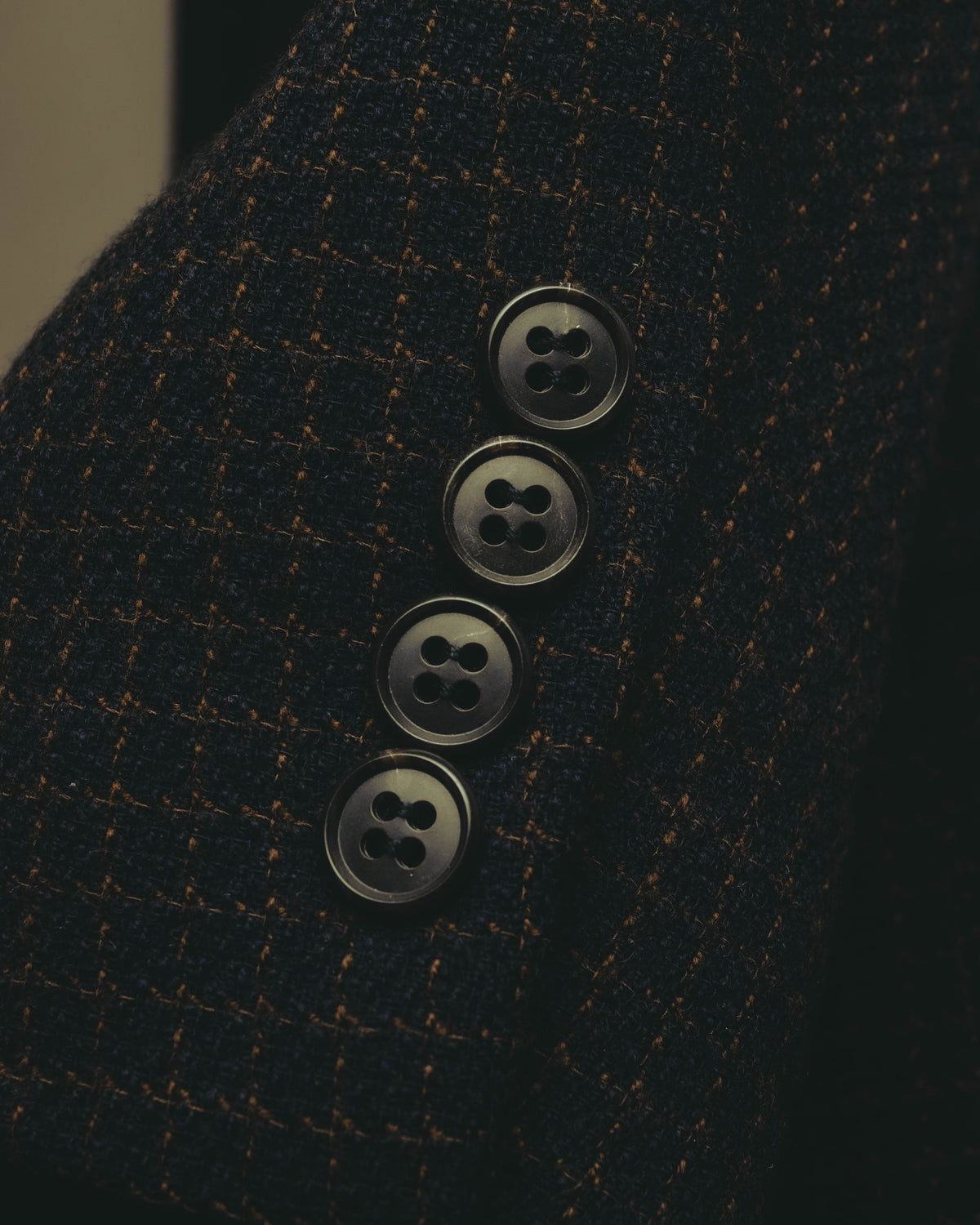 袖口に配された四つ穴ボタンと、ダークチャコールを基調にブラウン系の格子柄が重なる生地を捉えたディテールカット。細かな織りによる自然な陰影も確認できます。