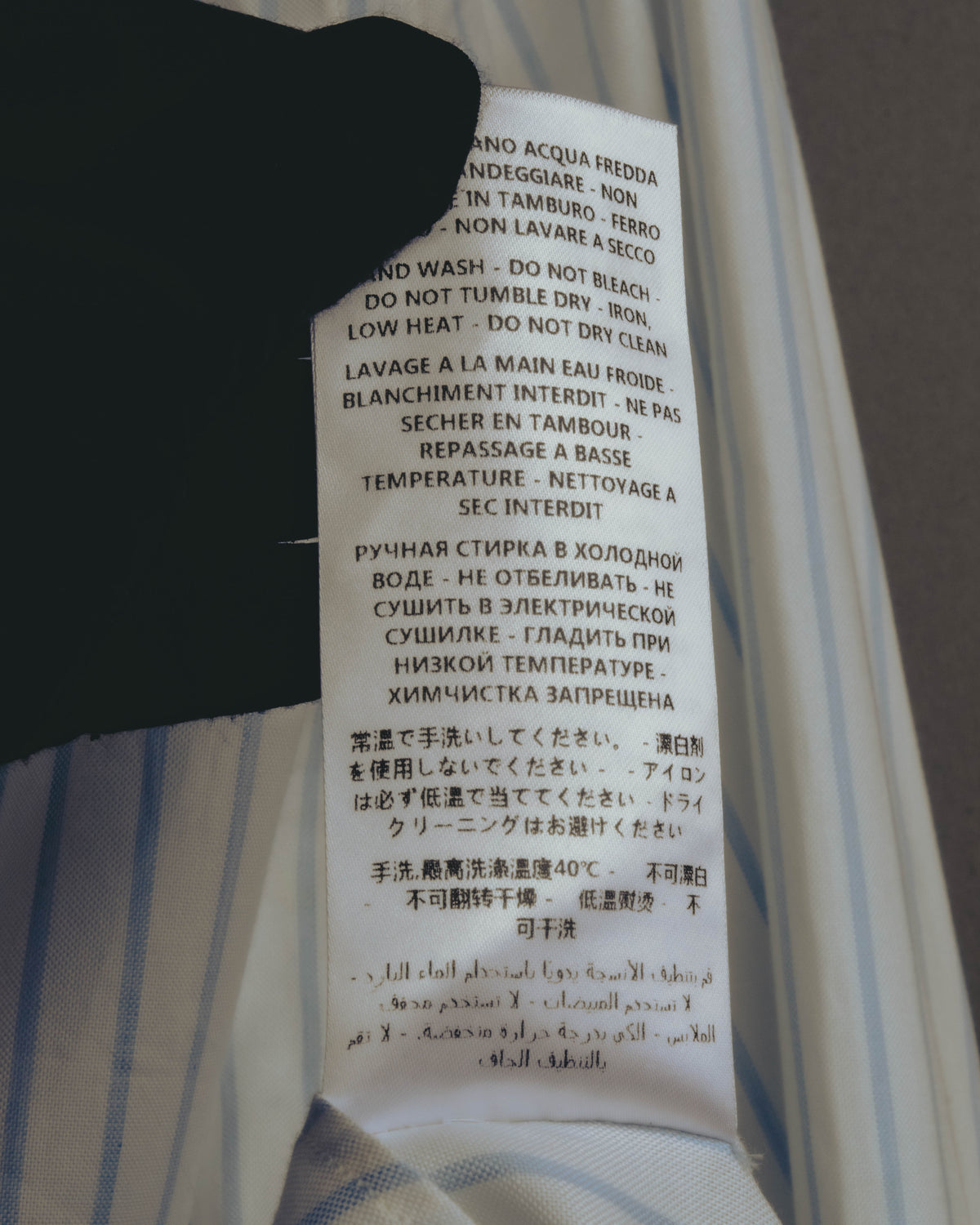 白地に淡いサックスブルーの細幅ストライプ生地に付された、多言語表記の品質表示タグ。手洗い・漂白不可・低温アイロンなどのケア表記を確認できるカット。