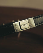 ブラックレザーストラップに配された、SAINT LAURENT PARISのロゴ刻印入りスクエア型メタル留め具（クラスプ）のアップ。直線的でミニマルなフォルムと、シルバーカラー金具の表情が確認できます。